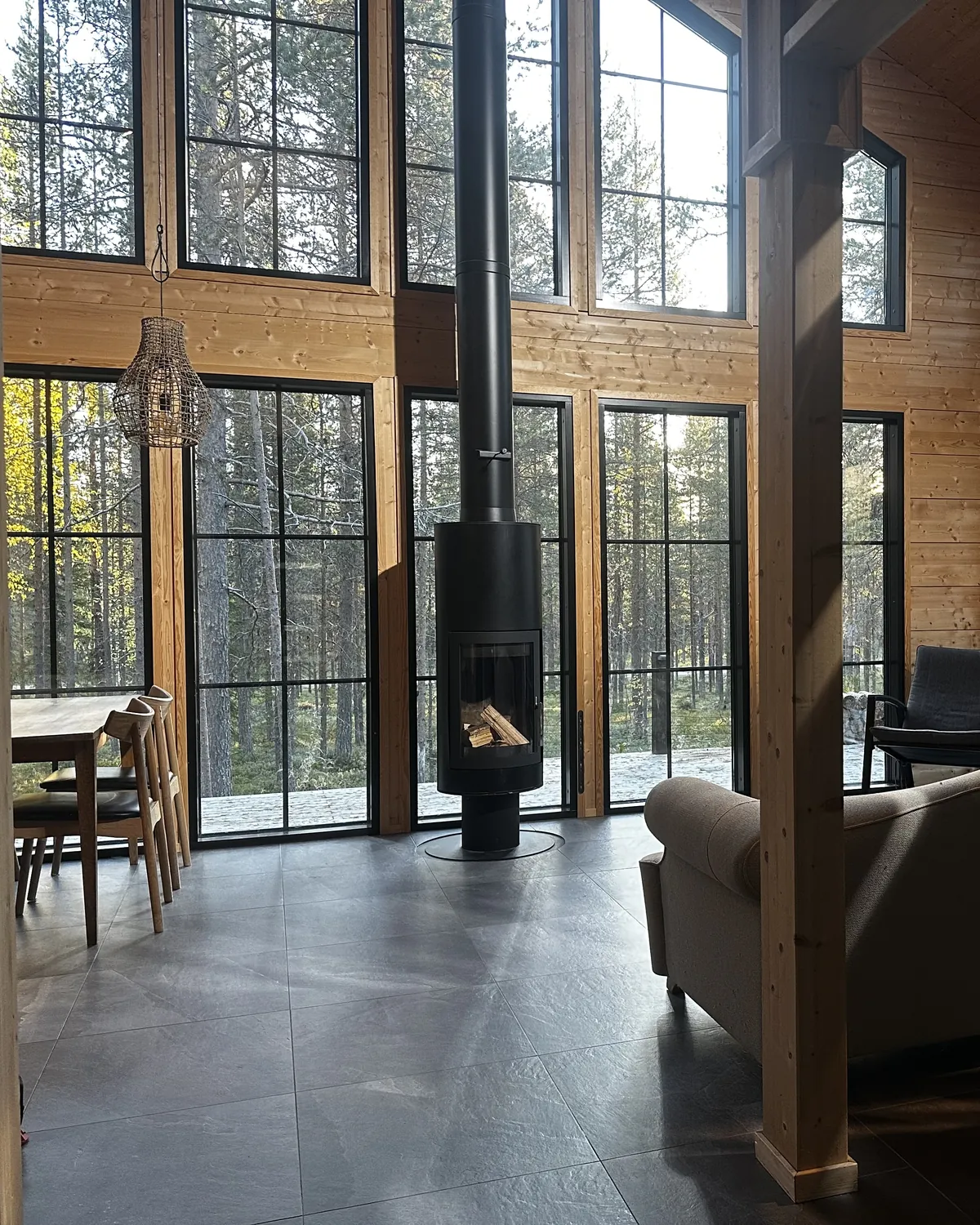 The double-height living area at Snow and Pine Log Cabin Levi — the central wood-burning stove framed by floor-to-ceiling glazing onto the forest